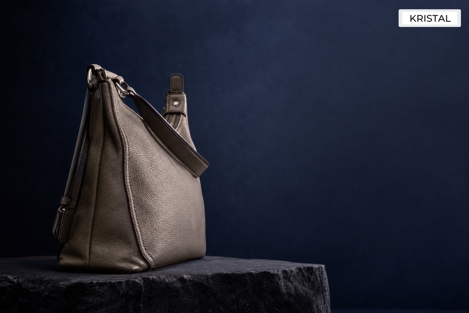 Side-profile luxury handbag on a dark stone plinth with ink-blue and silver light