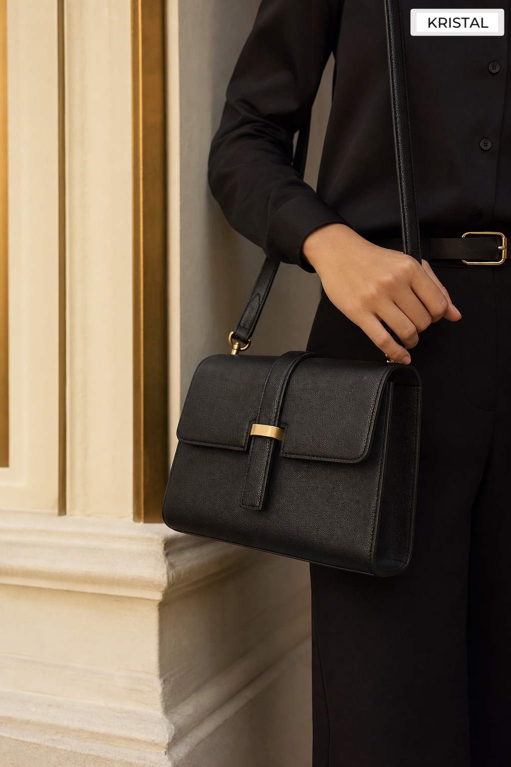 Black structured flap handbag held at waist height against an ivory architectural wall